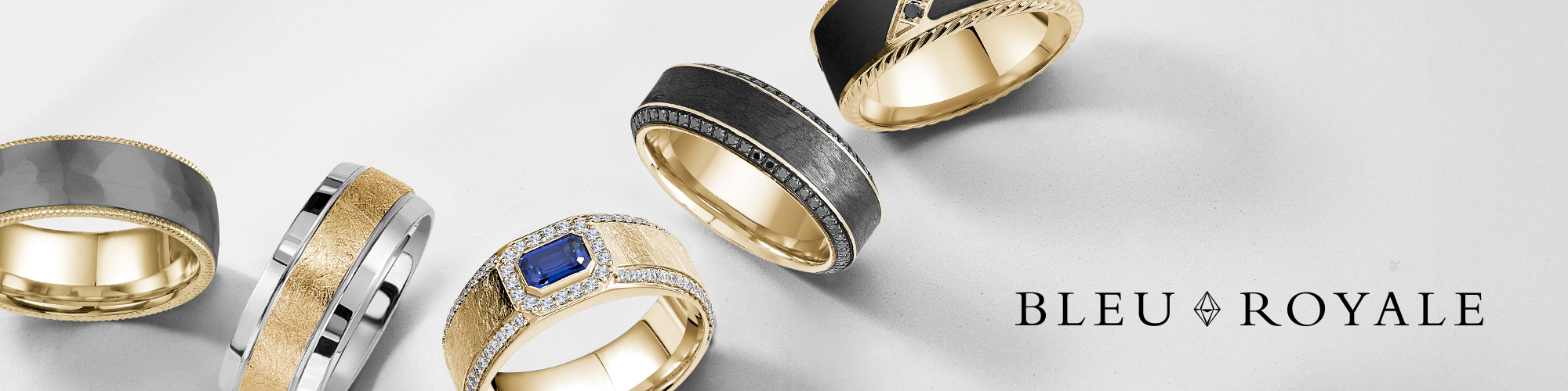 A stylish collection of five Bleu Royale men’s wedding bands displayed on a light gray surface. The rings showcase a variety of bold and modern designs, including combinations of gold, black, and silver tones, with finishes ranging from matte to polished. One standout ring features a vivid blue sapphire centerpiece surrounded by diamonds. The BLEU ROYALE logo is elegantly positioned in black text at the bottom right