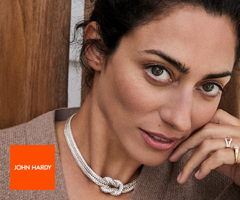 Close-up of a woman wearing a textured silver necklace with a knot design and a gold ring with a large gemstone, leaning her face on her hand. She is dressed in a brown top, and the John Hardy logo in a bright orange square appears in the bottom left corner