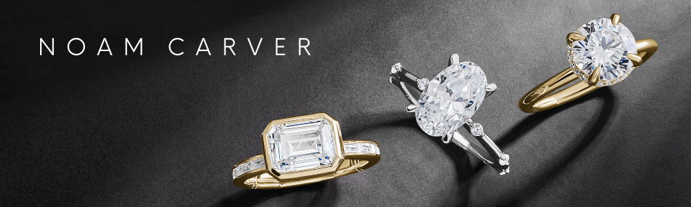 A luxurious display of three Noam Carver engagement rings set against a dark textured background. The rings feature large, brilliant-cut diamonds in different shapes and settings — a rectangular emerald-cut diamond in a yellow gold band with side stones, an oval diamond in a white gold setting with side baguette diamonds, and a classic round brilliant diamond in a yellow gold solitaire setting. The NOAM CARVER brand name is elegantly displayed in white text at the top left