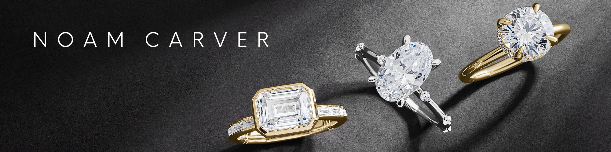 A luxurious display of three Noam Carver engagement rings set against a dark textured background. The rings feature large, brilliant-cut diamonds in different shapes and settings — a rectangular emerald-cut diamond in a yellow gold band with side stones, an oval diamond in a white gold setting with side baguette diamonds, and a classic round brilliant diamond in a yellow gold solitaire setting. The NOAM CARVER brand name is elegantly displayed in white text at the top left