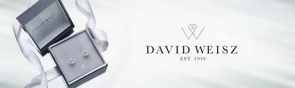 A pair of diamond stud earrings displayed in a grey velvet jewelry box with a matching lid embossed with the logo and name 'David Weisz EST. 1949.' A silver ribbon lies elegantly beside the box, emphasizing the luxury and sophistication of the presentation.