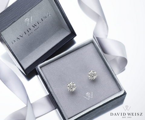 A pair of diamond stud earrings displayed in a grey velvet jewelry box with a matching lid embossed with the logo and name 'David Weisz EST. 1949.' A silver ribbon lies elegantly beside the box, emphasizing the luxury and sophistication of the presentation.