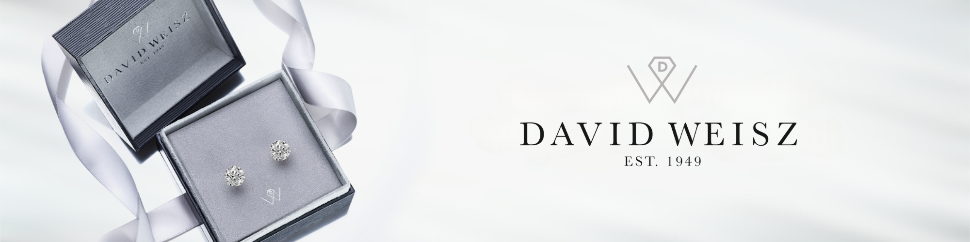 A pair of diamond stud earrings displayed in a grey velvet jewelry box with a matching lid embossed with the logo and name 'David Weisz EST. 1949.' A silver ribbon lies elegantly beside the box, emphasizing the luxury and sophistication of the presentation.