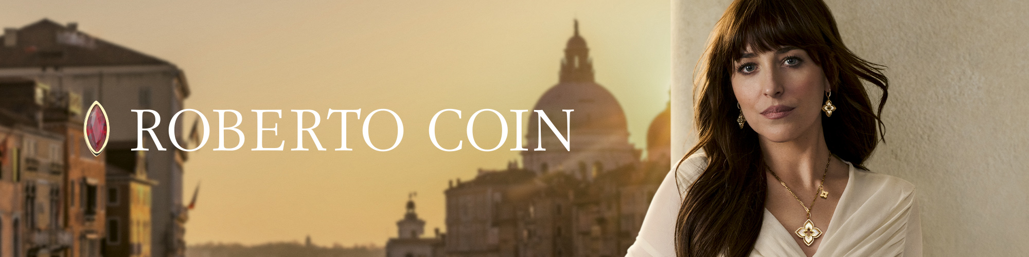 Actress Dakota Johnson dressed in an elegant cream-colored suit wearing gold Roberto Coin jewelry, seated in front of a sunset view of Venice, Italy. The text 'Roberto Coin' is displayed across the image