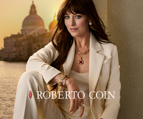 Actress Dakota Johnson dressed in an elegant cream-colored suit wearing gold Roberto Coin jewelry, seated in front of a sunset view of Venice, Italy. The text 'Roberto Coin' is displayed across the image